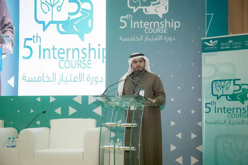 رجل مرتدياً الغترة والعقال والبشت السعودي عند منصة شفافة ليلقي كلمة خلال فعاليات الدورة الخامسة للامتياز الطبي في جامعة القصيم. اختر خلفه شعارات وشاشات كبيرة تحمل اسم الحدث باللغتين العربية والإنجليزية، مع تسليط الضوء على ورش العمل والمحاضرات الطبية المصاحبة.