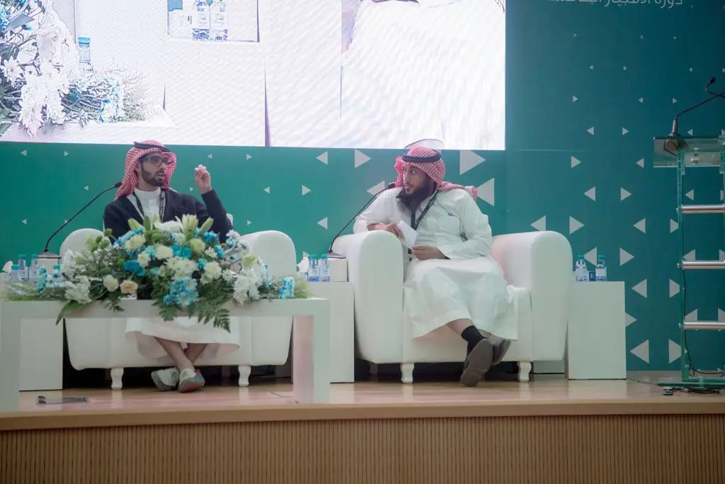 يرتدي رجلان الزي السعودي التقليدي، متمثلًا في الغترة أو الشماغ والعقال بالإضافة إلى البشت السعودي، على مقعدين أبيضين فوق المنصة في فعاليات دورة الامتياز الطبي في القصيم. يتبادلان الحديث في جو رسمي، بينما تتوسطهما باقة زهور. تذكر في الخلفية حامل ميكروفون وشاشة عرض كبير للفعالية المميزة.