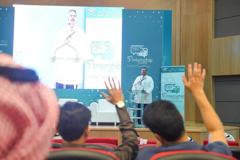 لا تعرف مرتديًا البشت السعودي والغترة والعقال على منصة الملقنين في قاعة المحاضرات، مخاطبًا الحضور الرسميين خلال فعاليات الفترة الخامسة الامتياز الطبي في القصيم. وفي الصفوف، رفع عدد من المشاركين الشماغ والسؤال طلبًا للتفاعل والاستفسار أثناء المحاضرات الطبية، بينما يرصد خلف الشاشة الكبيرة صورته بشكل واضح للحضور.