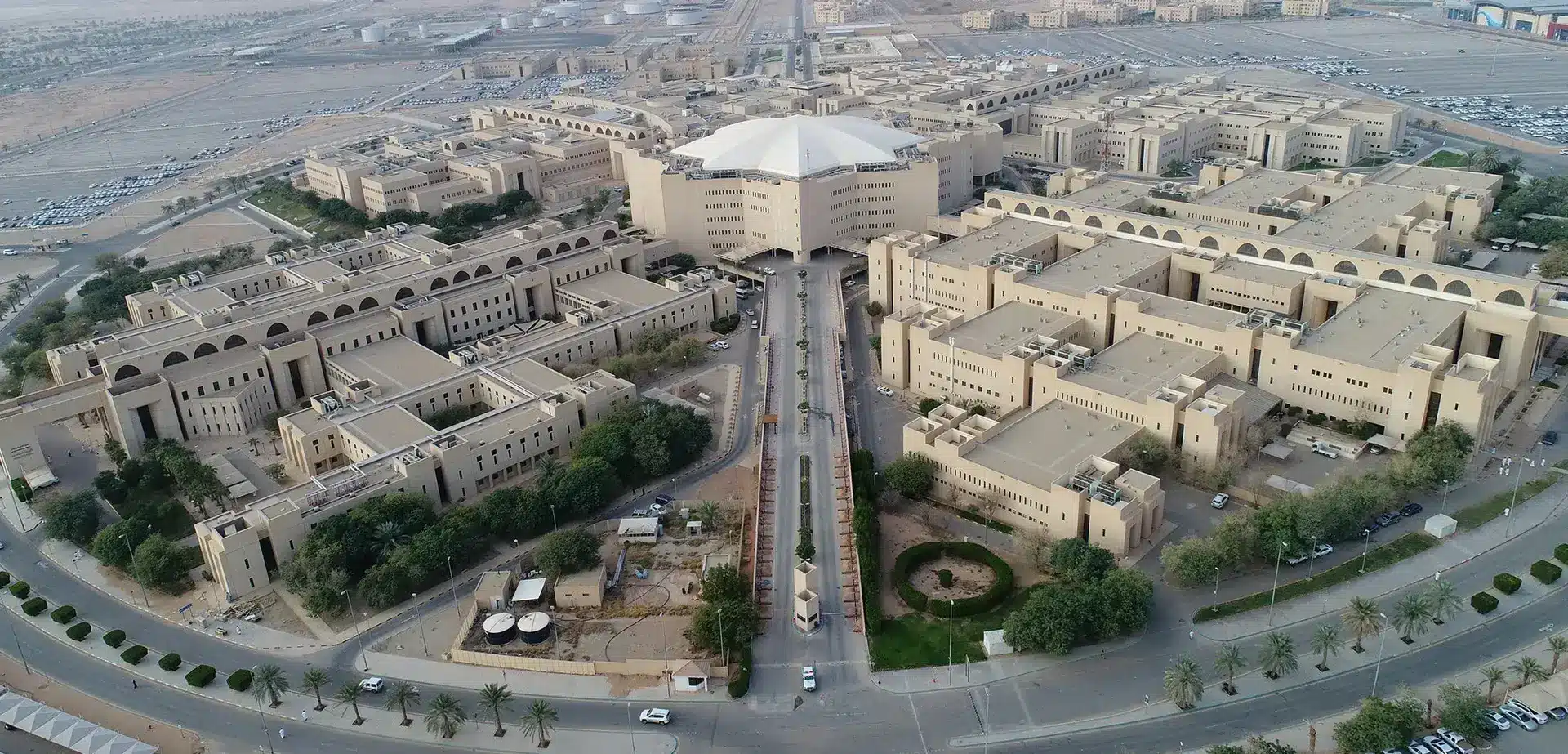 صورة جوية لجامعة القصيم الحرم الجامعي الحرم الجامعي الحديث بمبانيه حيث يوجد اللون الشامل، ويتسع وسطها مبنى رئيسي ذو سقف أبيض، وتحيط بها طرق مُظللة بالأشجار ومناطق مواقف السيارات مجهّزة خصيصًا لسيارات الطالبات. المشهد العام ينبض بالرقي ويتناسب مع البيئة النووية النووية، ويبرز الطلاب فيه والموظفون بارتدائهم الشماغ السعودي أو الغترة مع العقال والبشت السعودي في فعاليات رسمية، ًا على المؤكد الوطنية والهوية الثقافية للمملكة.