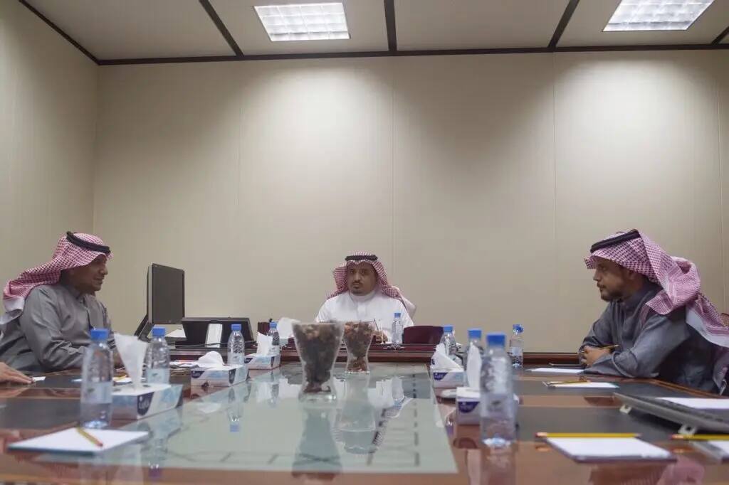 يجلس ثلاثة رجال على الغترة السعودية والشماع والعقال والبشتاء السعودي حول طاولة بدل في جامعة القصيم. توزع على جداول الأوراق والمستندات والقوارير، بالإضافة إلى شاشة حاسوب مخصصة للخدمات الإلكترونية. الاشتراك في غرفة الاجتماع نجحانها ذات اللون وإضاءة السقفية ساطعة لمسة رسمية للمكان.