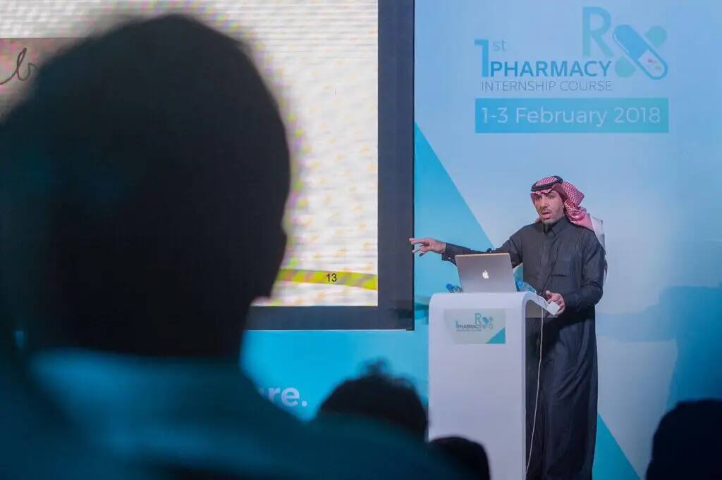 ارتدى رجل الغترة السعودية والعقال والبشت السعودي يقدم عرضًا جديدًا في كلية صيدلة في القصيم خلال فعاليات دورة العملاق، مشيرًا إلى شاشة خلفه تم الكشف عن براءة أول دورة امتياز في الفترة من 1 إلى 3 فبراير 2018.