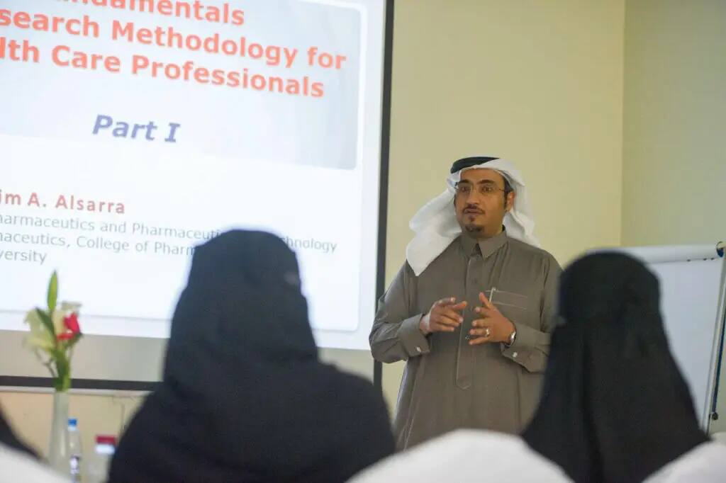 يرتدي رجل غترة بيضاء وقالاً مع بشت سعودي رسميًا، يقدّم محاضرة بعنوان "الأساسيات المطلوبة للبحث العلمي لتخصصي في الرعاية الصحية" لطلاب الامتياز من كلية الصيدلة في القصيم. حدقت في القاعة مزهرية زهرية وسبورة سوداء.