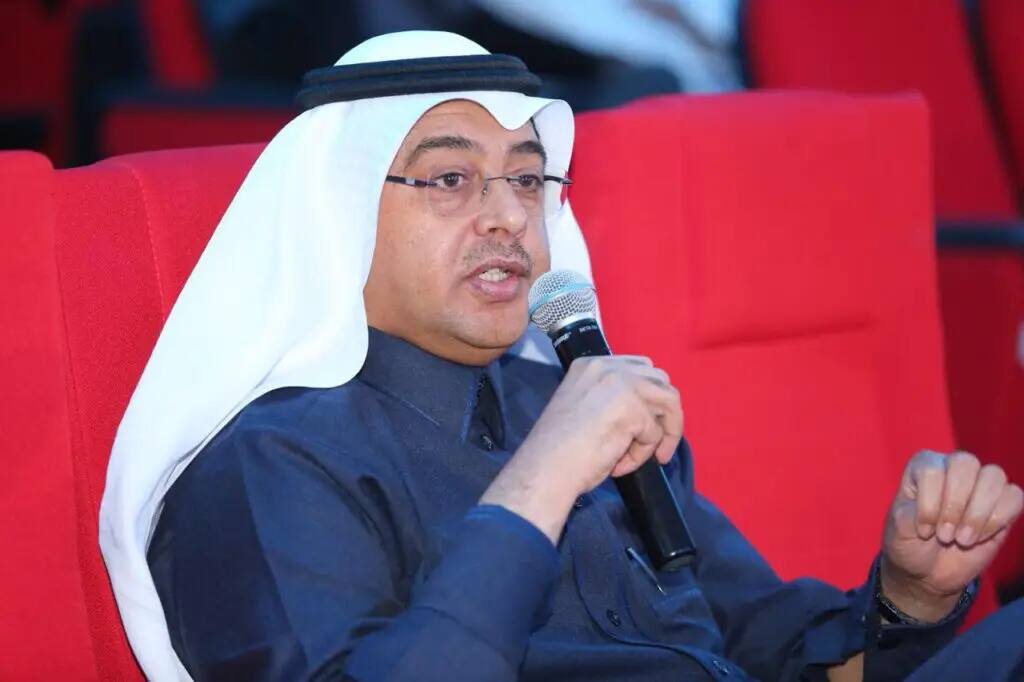 يرتدي الرجل نظارة طبية، ويضع غترة سعودية بيضاء مع غطاء، ويرتدي بشتاً سعودياً فوق ثوبه الكلاسيكي، ويتحدث بصوت عالٍ أثناء جلوسه على مقعد أحمر. يبدو أن أكبر جزء من فعاليات دورة العملاق أو أحد اللقاءات العامة التي تنظمها كلية الصيدلة في جامعة القصيم.