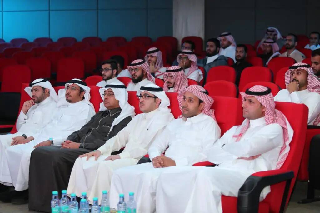 يجلسون مجموعة من الرجال مرتدين الغترة أو الشماغ والعقال والبشت السعودي، وفيهم عدد من طلاب العملاق من كلية الصيدلة، على مقاعد حمراء في قاعة المحاضرات، يوجهون اهتمامهم وفعالية دورة الامتياز. ساهم في تحضير موضوعة المياه في طاولة المقدمة.