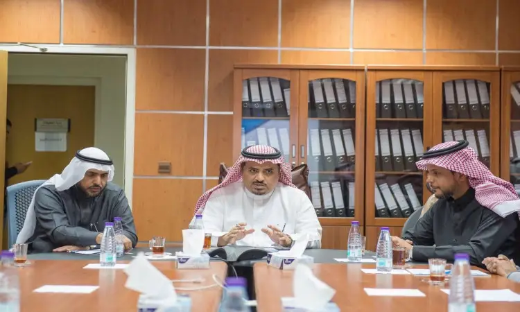 يجلس ثلاثة رجال على الغتة السعودية والعقال والبشت السعودي حول طاولة طعام في المكتب الرسمي، لذلك يستعرضون تقرير الإنجاز عن عمادة المعلومات التقنية. احتوت خلفهم رفوف مليئة بالمجلدات، في حين توجد على حسب الجدول الزمني للمياه ومناديل ورقية، مما يعكس المهنية والرسمية للاجتماع في جامعة القصيم.