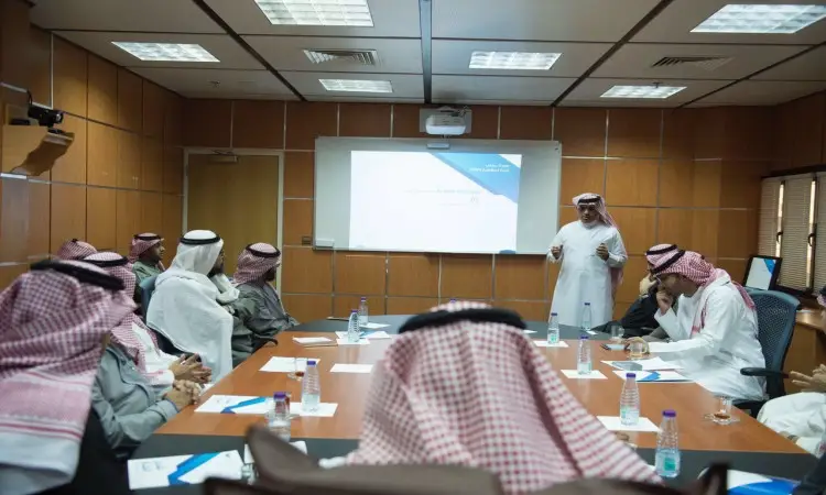 يجتمع عدد من الرجال مرتدين الغترة أو الشماغ السعودي مع العقال والبشت السعودي حيث يجلسون، حيث يستمعون باهتمام أحد مقدمي العرض الخشبي أمام شاشة تستعرض اشتراك اشتراك عن عمادة تقنية المعلومات، وذلك في قاعة المعيشة الفاخرة في القصيم.