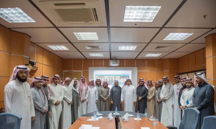 يجتمع مجموعة من الرجال تولى الغترة والشماع والعقال والبشت السعودي حول اجتماع في إحدى قاعات جامعة القصيم، حيث يلتقطون صورة جماعية مع متعة مدير الجامعة، بينما يُعرض تقرير الإنجازات الرائعة عن عمادة تقنية المعلومات على شاشة العرض في الخلفية.