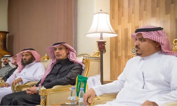 Three men dressed in the classic Saudi Arabian classical attire of the Ghutra or Saudi Shemagh with Eqbal, and the official Saudi Bisht, including the university director, sit on chairs inside a hall. In front of them is a table with two Saudi Arabian flags and bottles of water in the center, while the space is illuminated by a luxurious lamp and surrounded by wooden cladding. This immediate scene captures the essence of an ordinary Qassim University event.