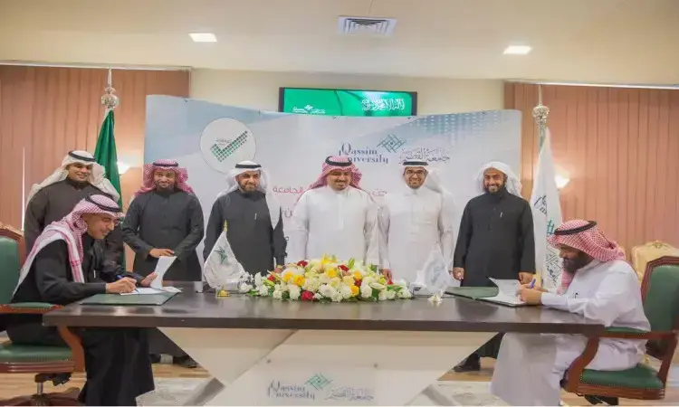 A group of men in formal Saudi dress, wearing the Saudi Ghutra or Shemagh, Eqal and Bisht, take a group photo as they sign the men's agreements at a table decorated with flowers at Qassim University. In the background are banners and flags bearing the logos of the university president and the Education Evaluation Commission.