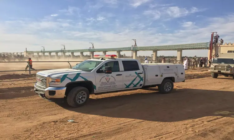 اللامعة للطوارئ، تحمل كتباً باللغة العربية، تتضمن "الجنادرية" و"مسيرة وإنجاز"، على طريق ترابي بالقرب من مجموعة من الأشخاص، وبعضهم يرتدي الشماغ السعودي أو الغترة مع العقال والبشت السعودي في مشهد يعكس الهوية الوطنية. وتظهر في خلفية الجسر تحت سماء صافية واضحة، بينما تتجلى جهود السلامة والتنظيم التي تتنافس بالتنسيق مع المحاكم، مثل جامعة القصيم.