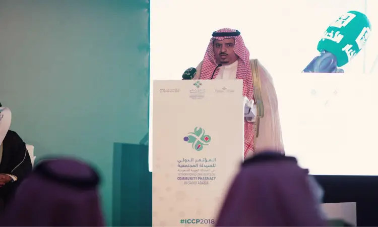 رجل يرتدي الزي السعودي الأصلي، شاملاً الغترة والعقال والبشت مايك السعودي، بوضوح على منصة المؤتمر الدولي للصيدلة، مستخدمًا ال بينما تُعرض صورته على شاشة كبيرة خلفه. اتخذ على المنصة شعارات المؤتمر والوسم #ICCP2018 مع إبراز وصيدلية المجتمعية.
