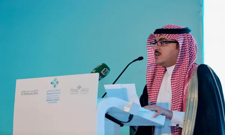 رجل القط مرتدياً الغترة السعودية والعقال والبشت السعودي خلف منصة بعدة ميكروفونات، ويحمل أوراقاً في يده. تسجيل تفاصيل المؤتمر الدولي للصيدلة المجتمعية باللغتين العربية والإنجليزية، وذلك ضمن فعاليات فعالية لجامعة القصيم على خلفية بالتركوازي.