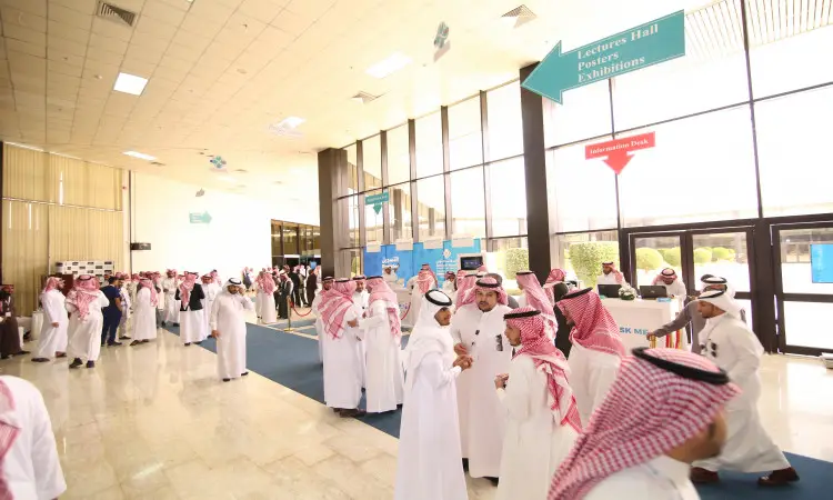 A large crowd dressed in Saudi Arabia's ghutra or shemagh, along with an aqqal and bisht, gathers on a wide, sun-drenched deck inside the Qassim Pharmaceutical Conference, where a blue banner directs attendees toward lecture halls, poster exhibits, and autograph signings. Some participants discuss pharmacy conference job opportunities, and the walk continues along the corridor.