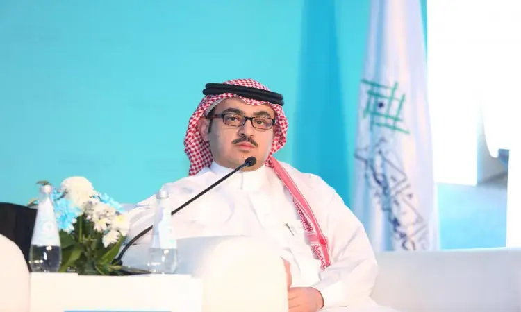 يجلس رجل يرتدي الغترة السعودية مع الشماغ والعقال والبشت السعودي على كرسي أبيض خلال مؤتمر الصيدلة المجتمعية في جامعة القصيم، وأمامه. ينصح في الخلفية بيضاء، وزجاجات مياه، بالإضافة إلى علم المملكة العربية السعودية.