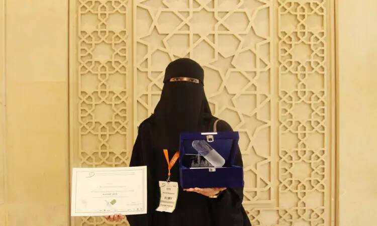 A Qassim University staff member, dressed in the Saudi Ghutra with Eqal and official Saudi Bisht, carrying a certificate and a blue box containing a glass shield, in front of an Arabic-themed ornament, proudly received the Dovat Award given to the university in recognition of its outstanding achievements during the Dubai International Pharmaceutical Technology Conference.