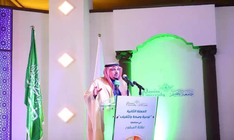 رجل يرتدي غترة وشماغقال وبشت سعودي للتواصل بشكل كبير على منصة غناء بميكروفون، بجانب علم المملكة العربية السعودية. تجدد خلفه شاشة البنفسج مضاءة بإضاءة خضراء، تحمل اسم "جامعة القصيم". تسلط الضوء على الجهود الجامعية في مجال التوعية والتثقيف الصحي بمحافظة عقلة الصقور.