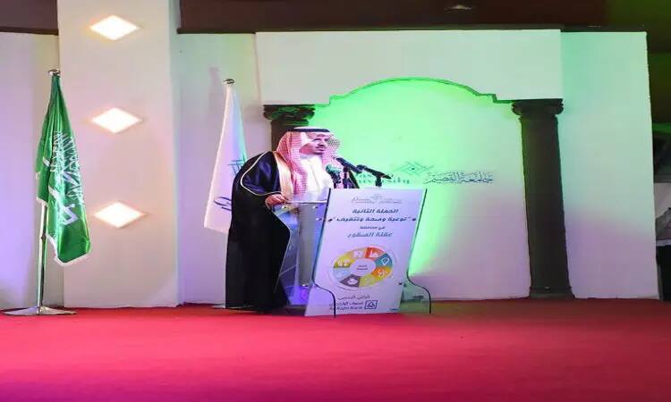 رجل مات مرتدياً غترة سعودية وقالاً مع شماع، وبشت سعودي، مشهوراً من على منصةٍ في قاعة مفروشة بسجاد أحمر. تتزين المنصة خلفه بإعلام المملكة العربية السعودية وأعلام منظمة. وتعرض المنصة شعار برنامج "توعية" خاتم الملوّن مع نص باللغة العربية. ويضيء الخلفية نورٌ أخضر في مقاطعة عقلة الصقور، وذلك ضمن فعالية المجتمع في جامعة القصيم.