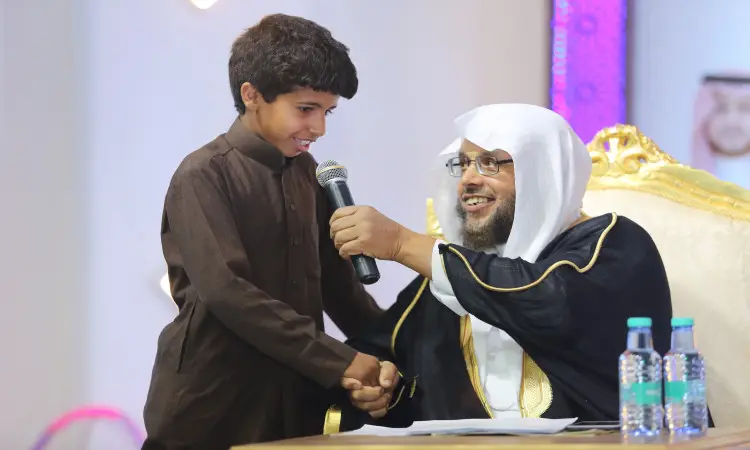 جلسة رجل بلباس سعودي تقليدي، يرتدي غترة وعقال وبشت، على كرسي ذهبي. ونجح في جامعة القصيم، قدم الميكروفون لطفل مبتسم لأطفاله على التايغا، ثم صافحه بحرارة. وقمت بإعداد مياه الشرب على الطاولة الأمامية.