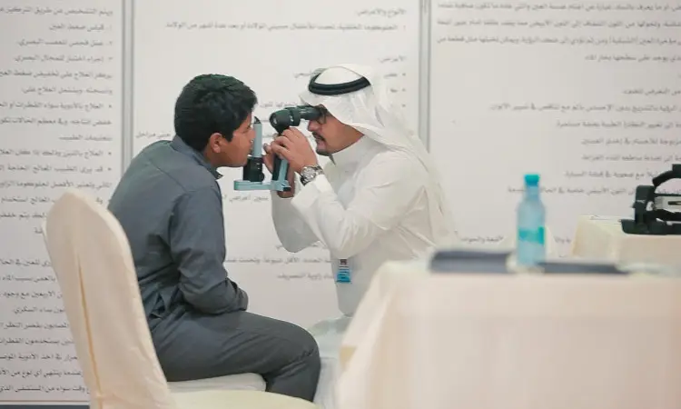 يرتدي رجل غترة سعوديةقالًا وبشتًا بشغل دوره في فحص العين لأحد الطلاب باستخدام الجهاز اليدوي في عيادة العيون في القصيم، وذلك بحضور عدد من الملصقات التوعوية باللغة العربية حول زراعة القرنية ومواضيع صحة العين الأخرى.