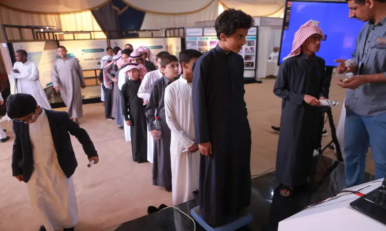 A group of young men wearing the Saudi Ghutra or Saudi Shemagh and Egal, as well as the Saudi Bisht, line up to participate in a health awareness event with a falcon mentality. One of the young men is seen stepping on the scale to help one of the supervisors. To see the number of attendees at the university events held inside the hall.