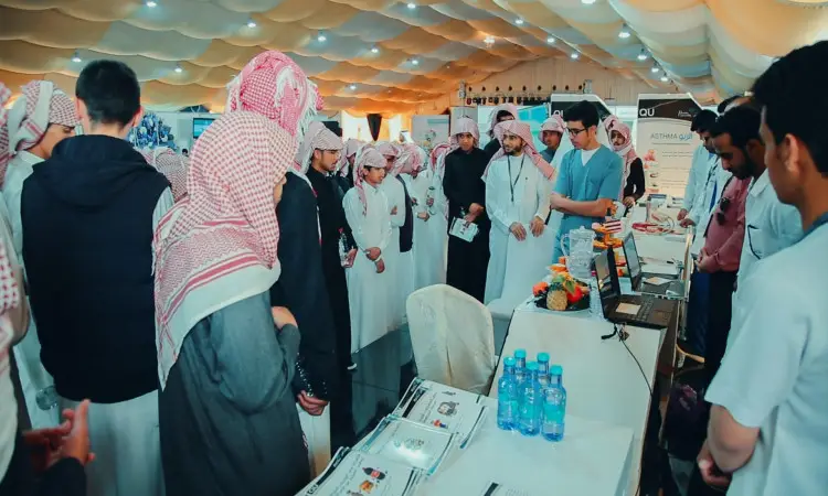 A number of men wearing the Saudi Ghutra or Shemagh, along with the Saudi Aqal and Bisht, gather at an indoor exhibition booth in Aqlat al-Suqur governorate, where Qassim University, wearing light blue shirts, is offering health awareness programs. Educational pamphlets, water bottles and some electronic devices are also on the tables.