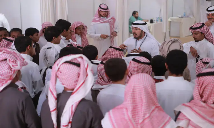 A group of young Saudi men and women, dressed in white robes and wearing Saudi shemaghs and aql, including an older-looking man in a Saudi bisht, are giving health education in a brightly lit hall. Two Lalan men in the background. This experiment is taking place in Aqlah al-Suqur province under the University of Qassim.