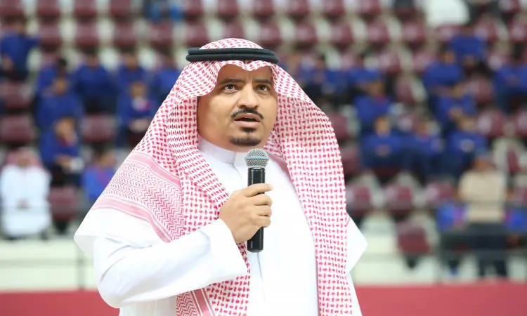 رجل غالباً ما يرتدي الغترة والعقال والبشتت السعودي عبر الميكروفون في نشاطات الهواء الطلق خلال بطولة الجامعات السعودية، بينما يحتل صفوف من الحضور الزي الأزرق وقد بدت صورتهم غير محددة.