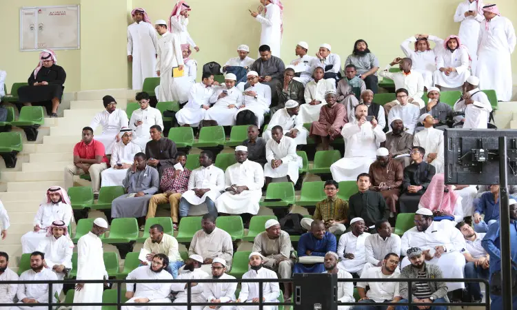 يجتمع عدد كبير من الرجال، وينظم العديد منهم الغترة أو الشماغ والعقال والبشت السعودي، على المدرجات الخضراء والبيضاء داخل الألعاب الرياضية في القصيم. يحضرون بانتباه وهم يتابعون فعالية غير ظاهرة في الصورة، ومن المحتمل أن يكونوا ضمن بطولة السعودية لكرة القدم. وبينهم ينهض للحديث أو استخدام الهاتف الجوال، بينما يظل يغلب عليه الطابعة الرسمية والقارئ الذي يعكس مكان الرؤية الصحيح.