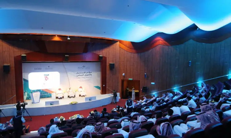 قاعة مؤتمرات في المملكة العربية السعودية، يجلس فيها الحضور على كراسٍ أحمر، ويرتدي العديد منهم الغترة أو الشماغ والعقال بالإضافة إلى البشت السعودي، بما في ذلك التوقيع التقليدي الرسمي للتمكن السعودي. يتواجد على المنصة عدد من الصحفيين يناقشون القضايا المتعلقة بالاستثمار والتمويل الصناعي وسط حضور إعلامي مكثف وعدد من الصحفيين الذين يسلطون الضوء على اهتمام الكبار.
