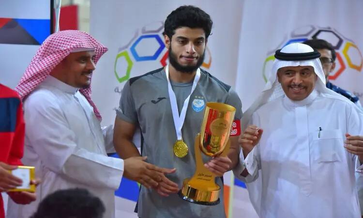 شاب رياضي يرتدي قميصًا رماديًا ممثلًا لمنتخب الجامعة، عصر بيده بطولة الكأس الذهبية ويرتدي ميدالية الفوز. رجل يحمل إلى جانبهان ويرتديتان الغترة والعقال والبشتت للرجال السعودي، يحيطانه بابتسامة فخر وعتزاز وبطولة الاتحاد الرياضي للصالات الرياضية.