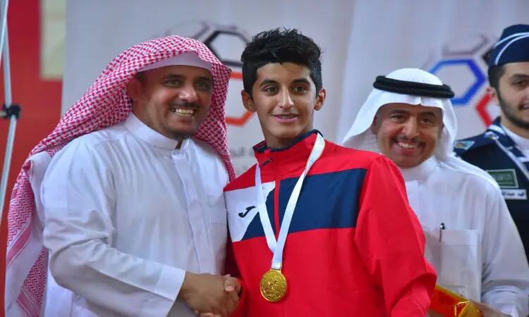 شاب رياضي من منتخب الجامعة، يرتدي ملابس رياضية حمراء اللون، يبتسم وهو وسط ميدالية ذهبية حول حاجبه أثناء مصافحته رجلاً يرتدي غترة وشماغاً وعقالاً مع بشت سعودي، وذلك خلال بطولة الاتحاد الرياضي، فيما بعد يبعث البهجة على وجوه الحضور في الخلفية.