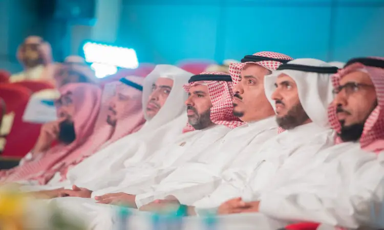 يجلسون عدد من الرجال مرتدين البشوت السعودية فوق أثوابهم البيضاء، وقد وضعوا الشماغات السعودية والأعِـقِلة بإتقان، على مقاعد حمراء في قاعة طبقات جامعة القصيم، متابعين باهتمام مراسم الكرام المتقاعدين.