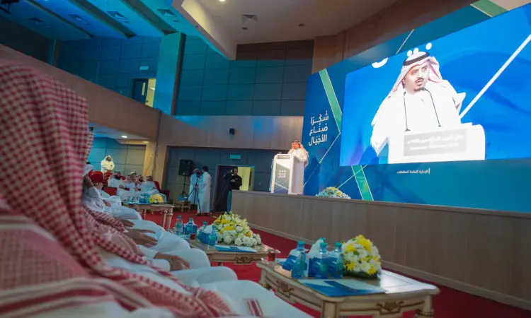 مطلوب رجل يرتدي الغترة السعودية والعقال والبشت السعودي كلمة من على المنصة خلال حفل تكريم المتقاعدين في جامعة القصيم، وقد عُرضت صورته على شاشة كبيرة خلفه. وحضرت القاعة الحديثة جمعا حديثا الذين ارتدوا الشماغ والعقال، وجُلِسوا بانضباط بين تنسيقات الزهور وقوارير الماء التي زينت المكان.