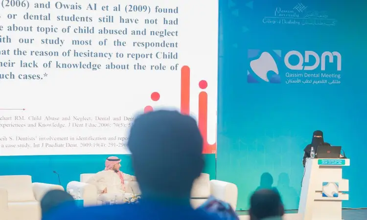 يُلقي كلمته من خلف منصة الخطابة خلال فعاليات ملتقى القصيم لطب الأسنان، المُقام في مركز الملك خالد الحضاري، حصر خلفه شاشة كبيرة حيث تم نصًا بحثًا ذا أهمية علمية. لعد عدد من الضيوف مرتدين الشماغترة والعقال والبشتت السعودي الرسمي، وينتابهم اهتمام بالغ بما بما يُعرض. وتبرز الهوية البصرية للفعالية ضدها الزرقاء والبيضاء المشرقة في أرجوة القاعة، ضمن احترام السيدات مكانة جامعة القصيم الفضيلة.
