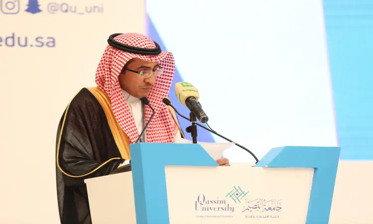 رجل يرتدي غترة وشماغقال وبشت سعودي خلف منصة الخطابة التي تحمل شعار جامعة القصيم، عظيمًا خلال مؤتمر الخصوصة وهو يقوم بكتابة بيده. ويتواجد أمامه ميكروفونٌ بغطاء أخضر، بينما يلتقط النصوص الخلفية باللغة العربية والإنجليزية.