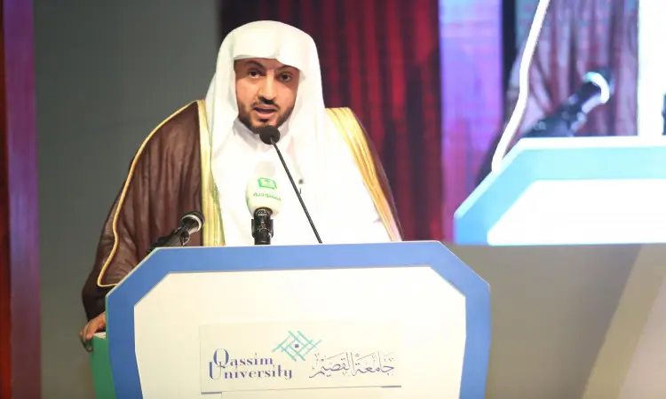 رجل يرتدي غترة سعودية وعقالًا وبشتًا سعوديًا يلقي كلمة من على منصة تحمل اسم "جامعة القصيم"، وأمامه عدد من المكروفونات، وذلك من خلال مؤتمر مؤتمر المجتمعوة المقام في القاعة الداخلية، بحضور سمو أمير منطقة القصيم.