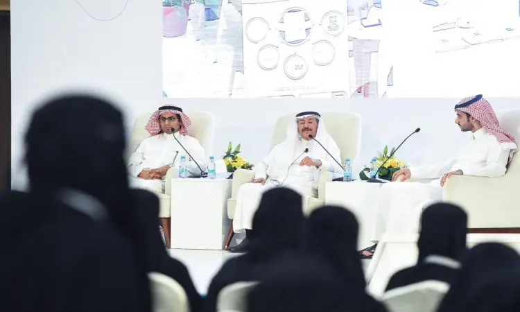 ضبط ثلاثة رجال ينتظرون الغترة أو الشماغ والعقال والبشت السعودي على منصة الفعاليات خلال فعاليات "دردشات النجاح"، حيث يشاركون في ندوة نقاشية. ويستمع للحضور، ومن بينهم نساء بملابس بيضاء، بينما تعرضن خلفين الشاشة الرئيسية خلال فعالية "يوم المهنة" المقامة في جامعة القصيم.
