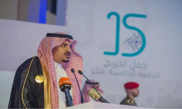 رجل مرتديا الثوب السعودي مع شماغ وعقال وبشت سعودي، المتحدث الكبير من على منصة متحكمة بالميكروفونات. يظهر خلفه شعار جامعة القصيم ونص عربي بارز، فيما يتعلق بعدد كبير من رجال الأمن بزيهم الرسمي في الخلفية.