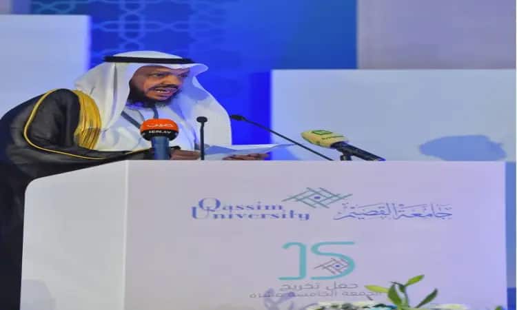 يتفضل رجل مرتدياً غترة سعودية وقالاً وبشتاً سعودياً، بالحديث من على منصة تدرب بميكروفونات خلال التخرج من جامعة القصيم، محترقة بكل خريج وخريجة. وتبرز على المنصة شعار الجامعة ونصوص عربية.