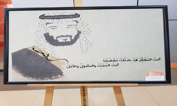 تُعرض لوحة مؤطرة على شماع، ويظهر فيها بورتريه مطرزة لرجل يرتدي الغترة والعقال والبشت السعودي التقليدي، إلى جانب الجانب العربي ويبرز اسم كلية التصاميم. وتأتي خلفية بيج فاتح، مع بطاقة صغيرة مُثبتة في الجزء السفلي من الإطار.