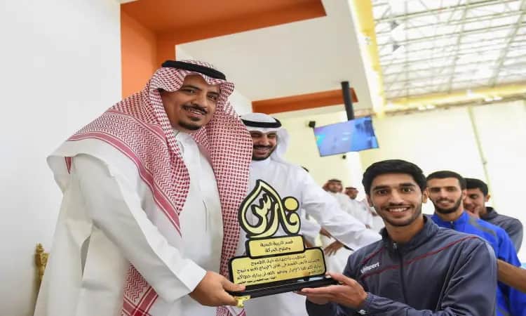 A man dressed in a Saudi Arabian ghutra, headband and bisht handed the "Fares" award to a smiling young man dressed in sportswear. to a smiling young man dressed in sportswear, in honor of his achievements in the field of kinesiology, amid a crowd of onlookers and a television screen in the background.