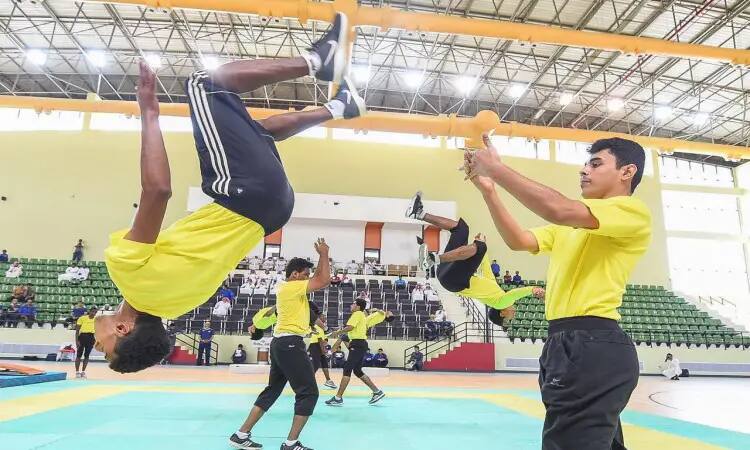 A group of people dressed in shirts with Saudi Arabia's Ghutra, Aqal and Besht shirts perform aerobatic movements and show jumps inside a gymnasium furnished with rugs for trainees, where they are unaffected by the science of movement under a pressure era coach who beats them. A number of attendees sit in the sports stadium to follow these exercises in the Qassim University's academic stature.