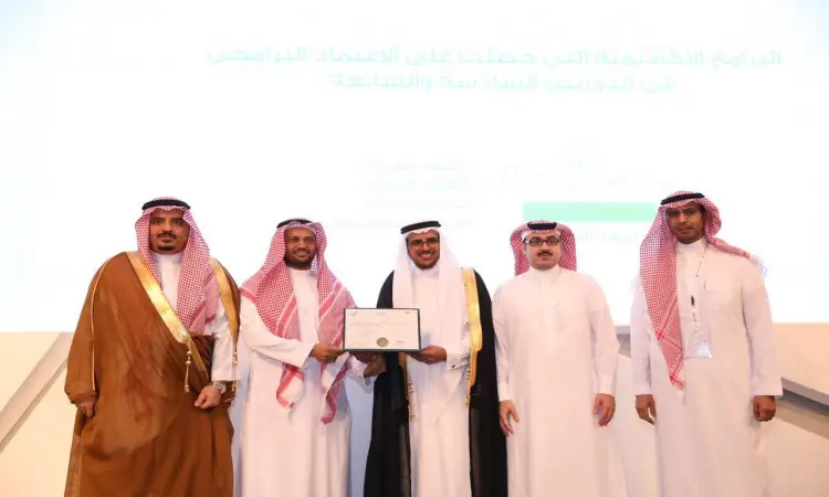Five men dressed in Ghutrah or Shemagh with Eqal and Saudi Bisht on stage. The men have worked in the university setting and are striving for academic accreditation from the National Assessment Center. Behind them is a display screen with texts in Arabic, from a center in Qassim.