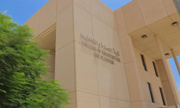 A colorful beige building with the words "College of Architecture and Planning" and "College of Architecture" written in English and Arabic, with a fresh green tree on the left side and a clear blue sky above the picture. At a time when Qassim University welcomes its distinguished students, the community wore the Ghutra or Shemagh and Egal with the Saudi Bisht, as part of the field training or study abroad program (Malaysia), as the scene embodies the spirit of belonging and academic excellence of the university.