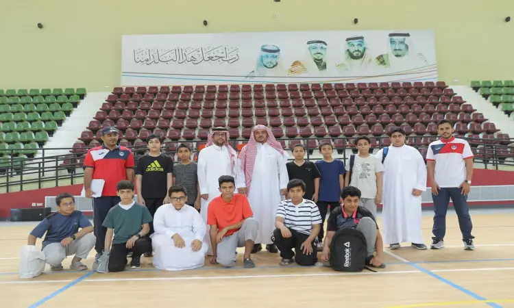 A group of students and men, including local students, pose for a group photo in a gymnasium, with a green and red base. The backdrop of the photo features prominent portraits and Arabic texts about the summer club and its activities. Some of the participants wore sports uniforms, while Cuba appeared in traditional Saudi dress, including the Saudi shemagh or Saudi ghutra with the Saudi aqal and bisht, including, of course, the official one at Qassim University.