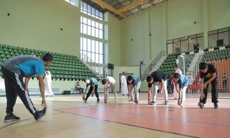 A group of students from the region, dressed in the Saudi Ghutra or Saudi Shemagh, Egal and Bisht, participate in back-to-back stretching exercises by touching their toenails inside the gymnasium, which is decorated with green seats, high windows and the Pope's goal with nets. A trainer supervises their execution of the exercises as part of the programs and activities of the summer club hosted by Qassim University.