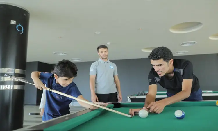 A young boy accurately shoots billiards on the table, supervised by a man wearing a Saudi jacket, headband and bisht, while a student watches the final scene in a modern indoor hallway as part of a summer club for students in the region, organized by Qassim University.