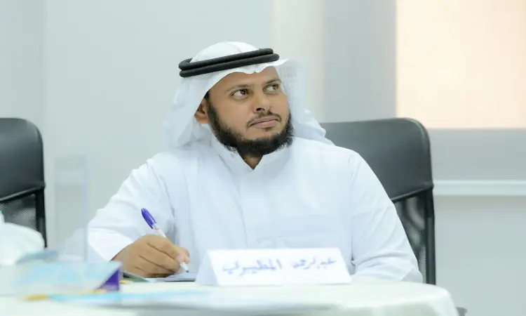يرتدي الرجل زيًا أبيض، وغترة سعودية مع عقال وبشت سعودي عند طاولة الاجتماع، متجهًا بنظره إلى الجانب أثناء الاجتماع الجامعي حول التطوير المهني للمعلمين في جامعة القصيم. تتواجد على جدول لوحة اسمية وأوراق تخص دورة المقررات.