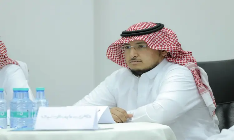 يرتدي رجل يرتدي اللباس السعودي التقليدي، يشمل الغترة السعودية البيضاء ويا الشماغ الأحمر الأبيض مع العقال، بالإضافة إلى البشت السعودي باختيار، على طاولة يتصدرها على لوحة تعريفية وبجوارها التعاونية مياه. يشارك الرجل في الاهتمام الاجتماعي ضمن دورة العمل أو برنامج التطوير المهني للمعلمين في جامعة القصيم.
